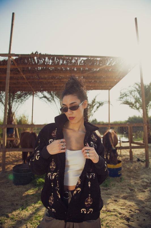 A woman standing outdoors wearing sunglasses and a black jacket with embroidered patterns. There are horses in the background.