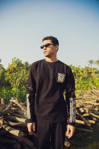 A person standing in a natural outdoor setting with wooden logs in the background, wearing a black long-sleeve shirt with gold embroidery on the lower sleeves and sunglasses.