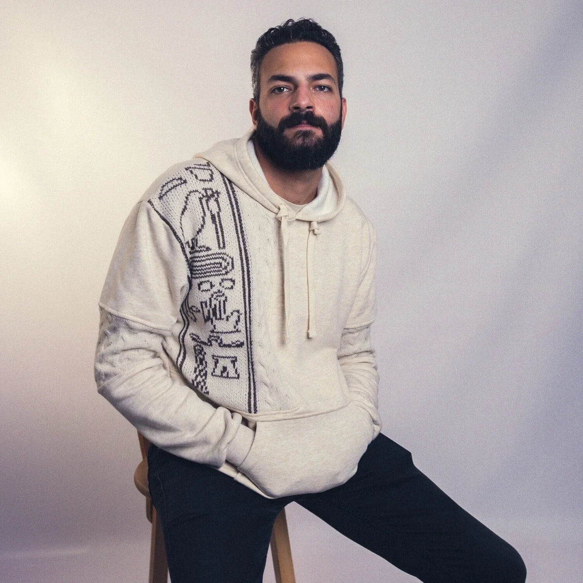 Man wearing beige hieroglyph hoodie, seated studio portrait