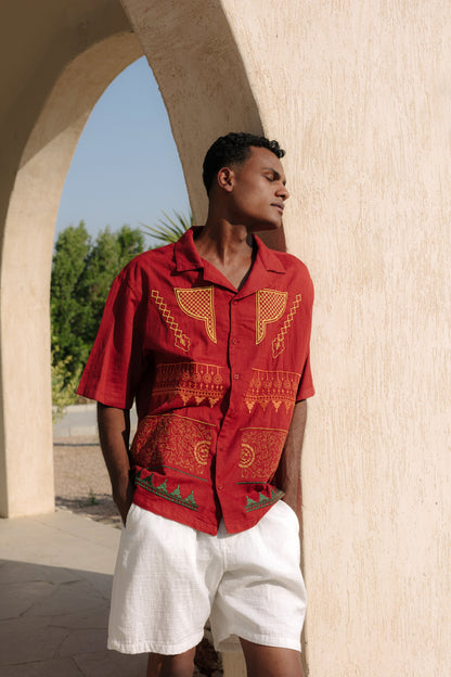Man wearing a maroon embroidered nubian shirt and white shorts standing under an archway.