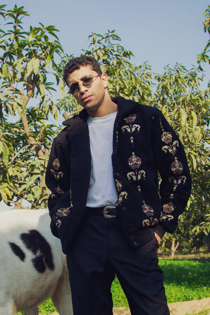 A person wearing a black and beige checkered jacket with embroidered floral patterns, paired with a white shirt, standing outdoors with trees and a cow in the background.