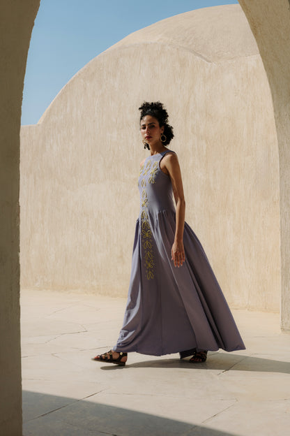 Woman in a long, light purple dress standing in front of a beige wall with a blue sky.