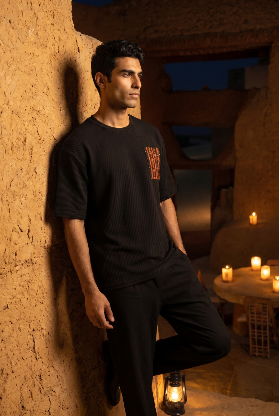 A man in a black T-shirt with a stitched pocket, leaning on a mudbrick wall at night