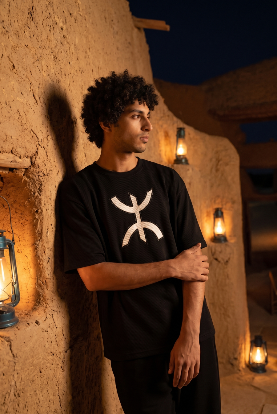 A man in a black T-shirt with a white Amazigh symbol, against a lantern-lit mudbrick wall