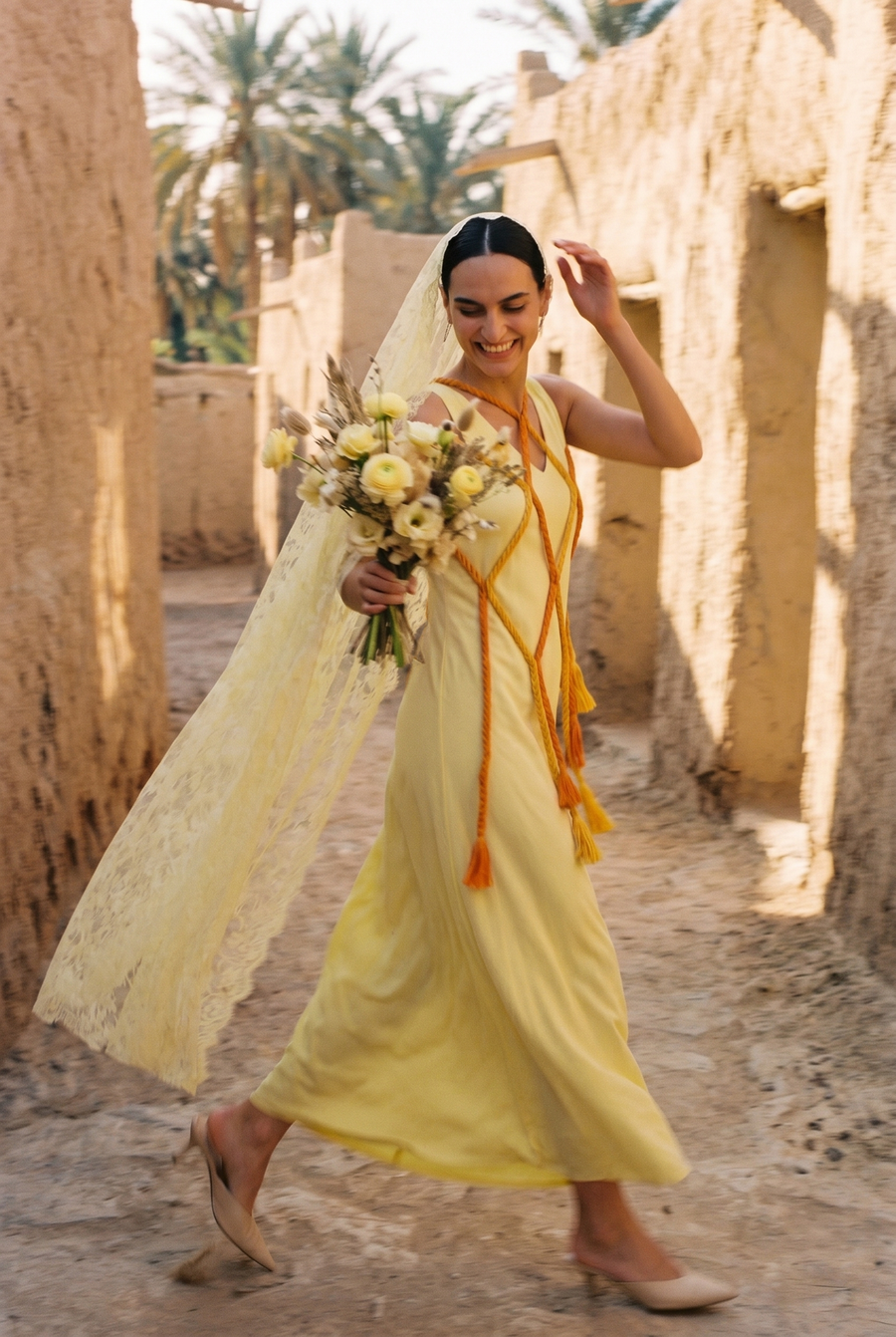 A woman in a yellow dress with orange rope ties and a lace veil walks through a mudbrick lane, holding yellow flowers
