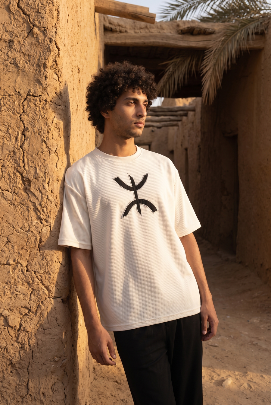 A man in a white T-shirt with a black Amazigh symbol, leaning on a mudbrick wall