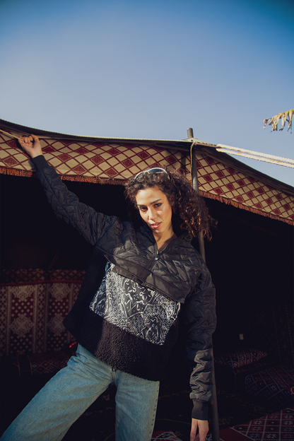 A person wearing a black sweatshirt with a geometric pattern, posing with one arm raised against a backdrop of a traditional structure.